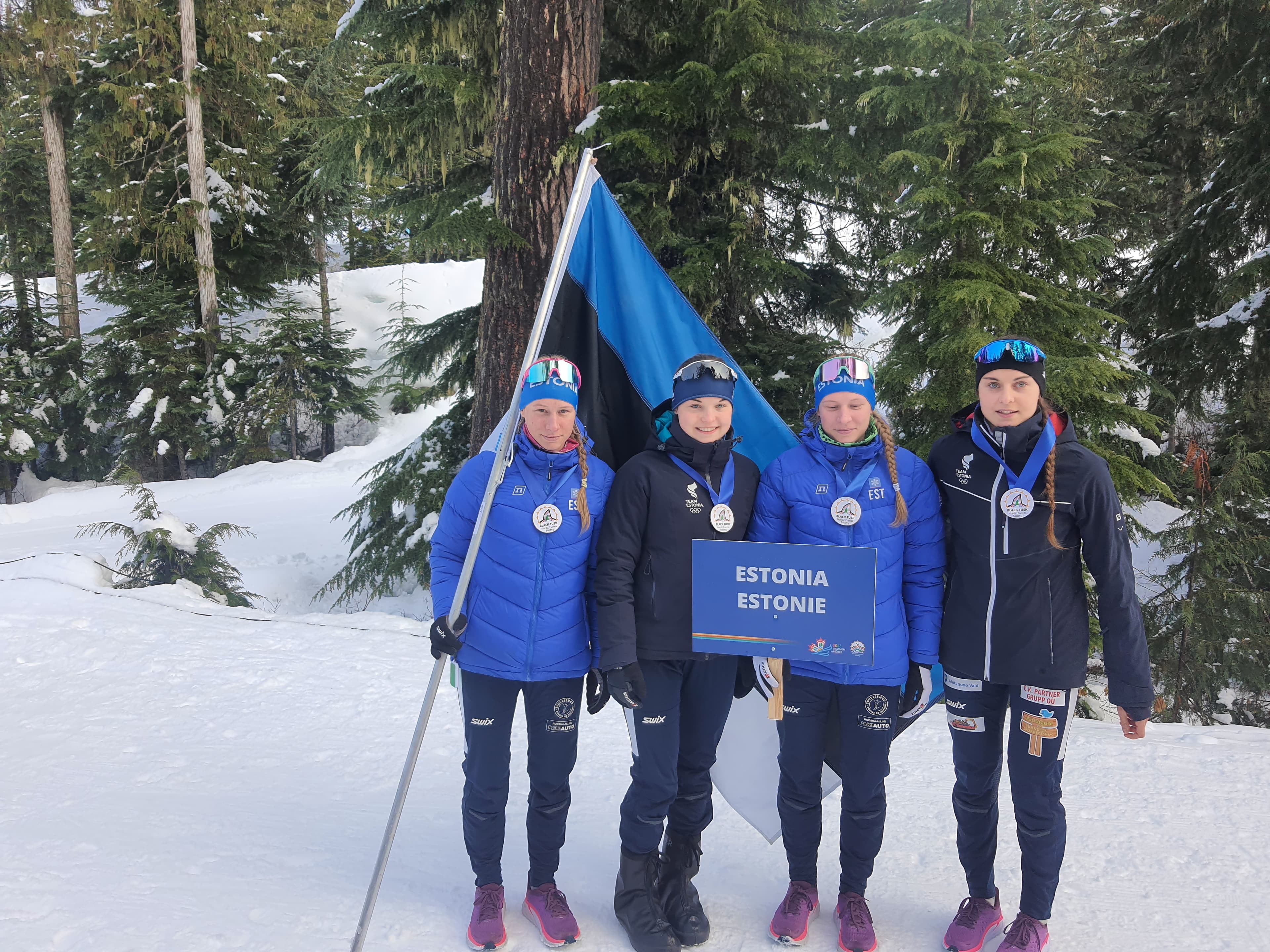 Four Estonian female athletes pose with medals and flag in a snowy forest setting.