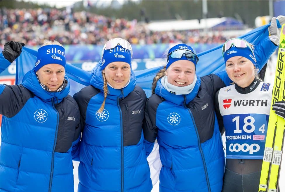 Four female Estonian cross-country skiers in blue uniforms pose together holding a blue flag.