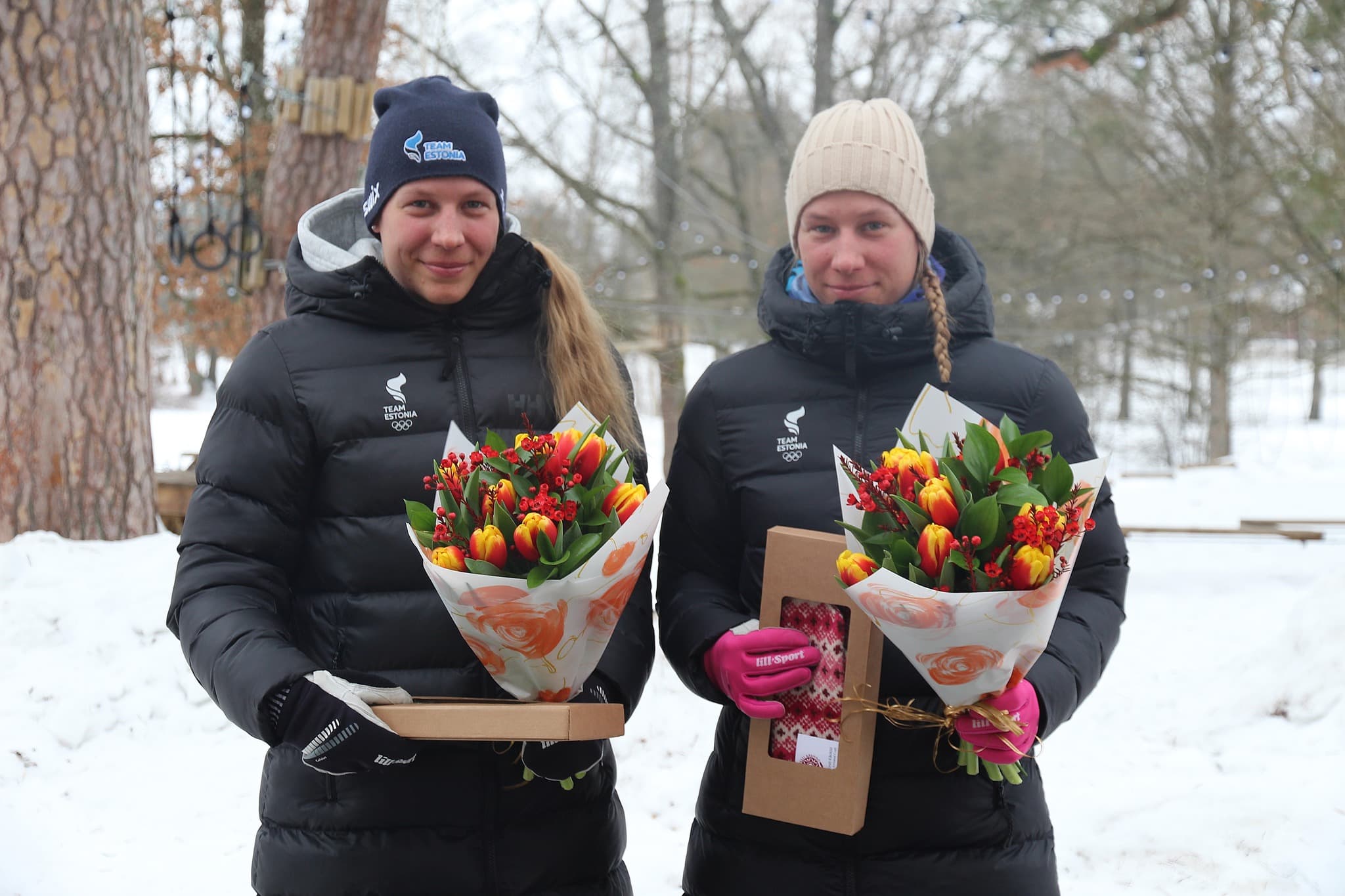 Two Team Estonia athletes in black puffer jackets hold tulip bouquets in a snowy park.