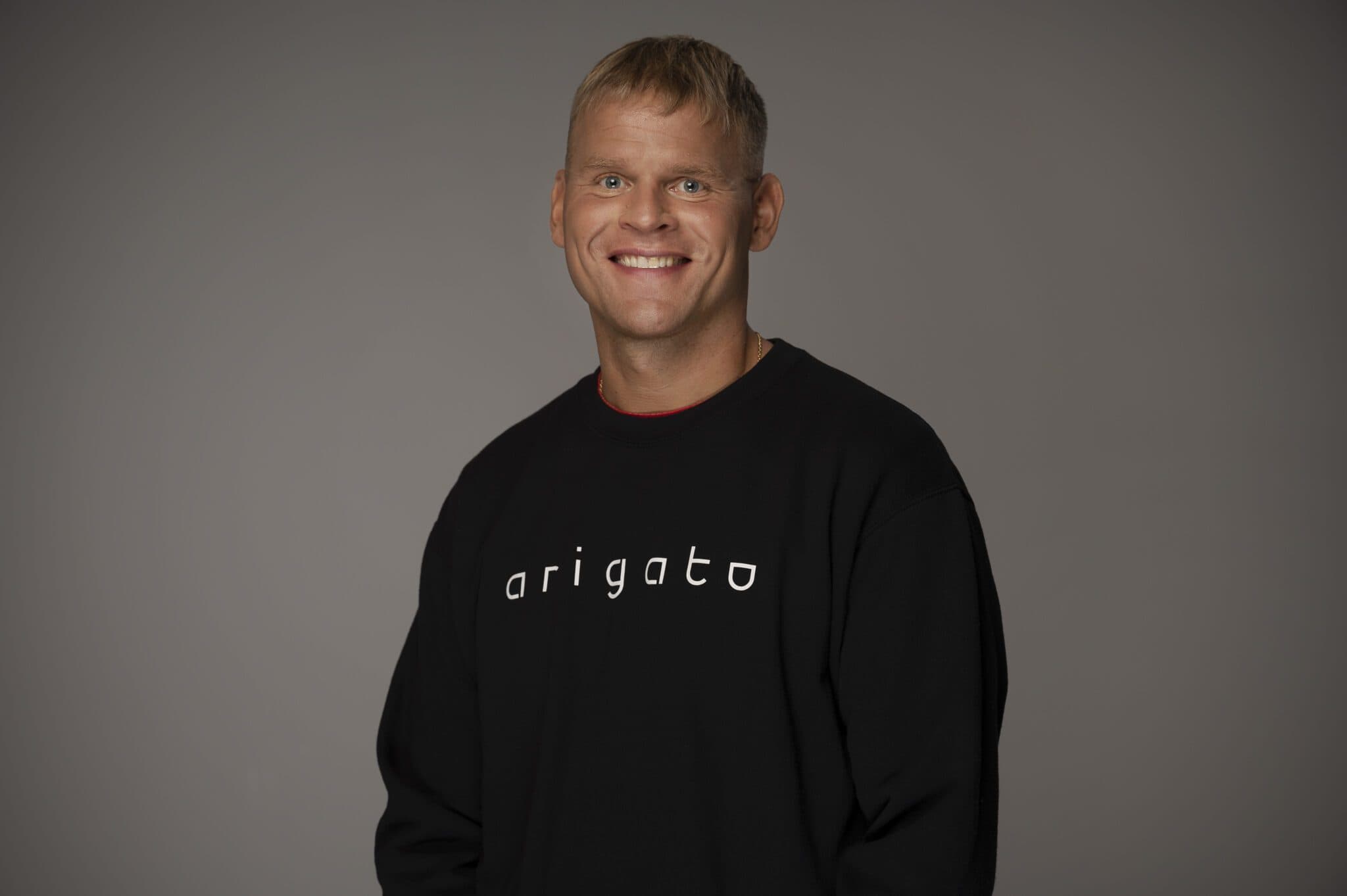 Smiling man with short blond hair wearing a black 'arigato' sweatshirt against grey background.