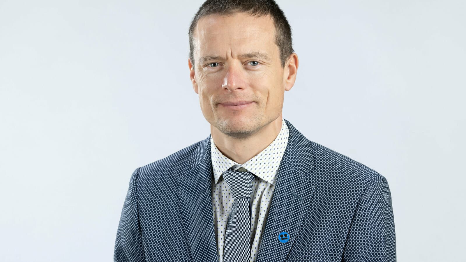 Middle-aged man with short hair wearing a blue patterned blazer and a grey patterned tie.