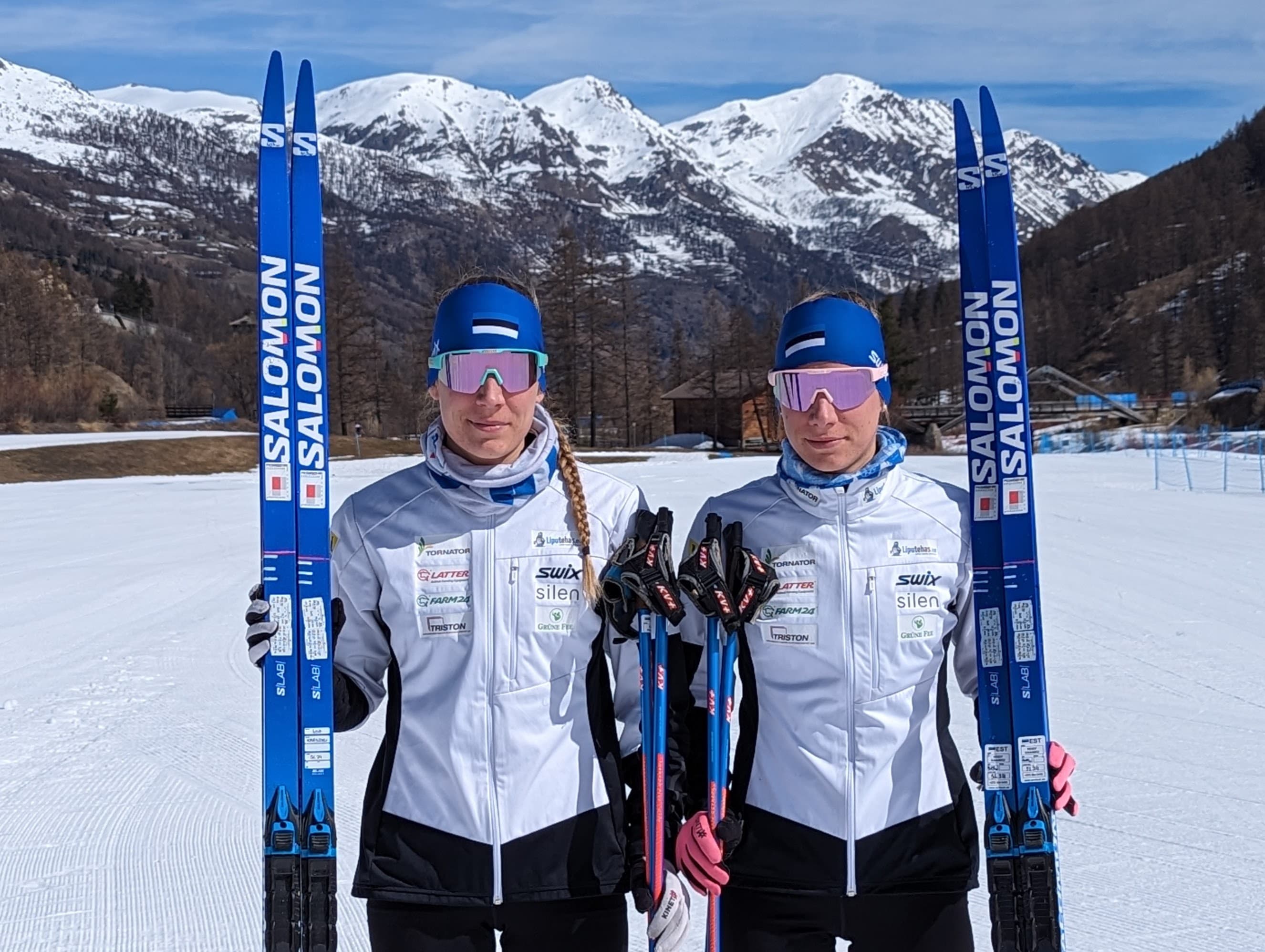 Two female Estonian cross-country skiers holding blue skis in a snowy mountain landscape.