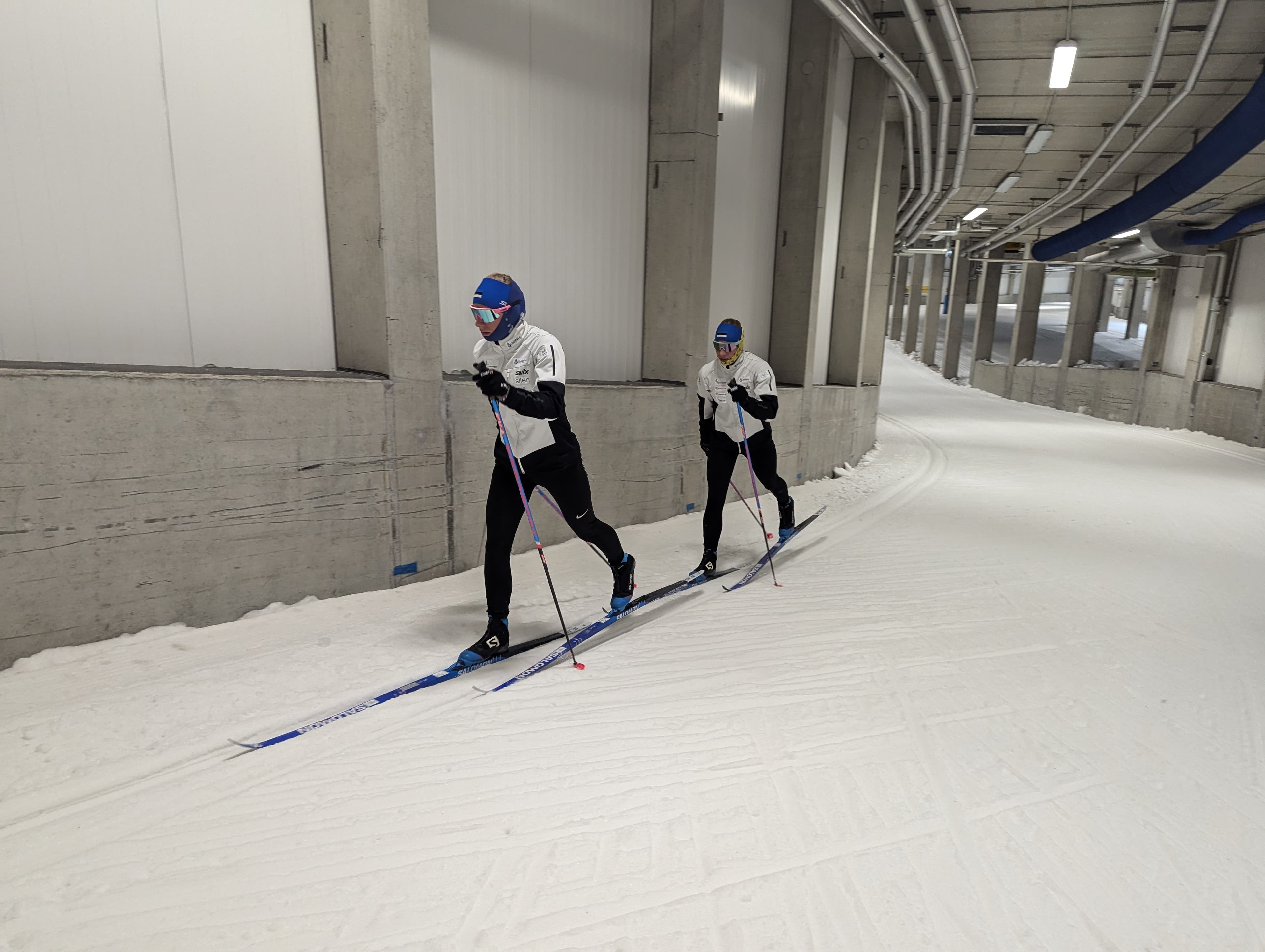 Two cross-country skiers glide along a groomed track inside a curved concrete indoor tunnel.