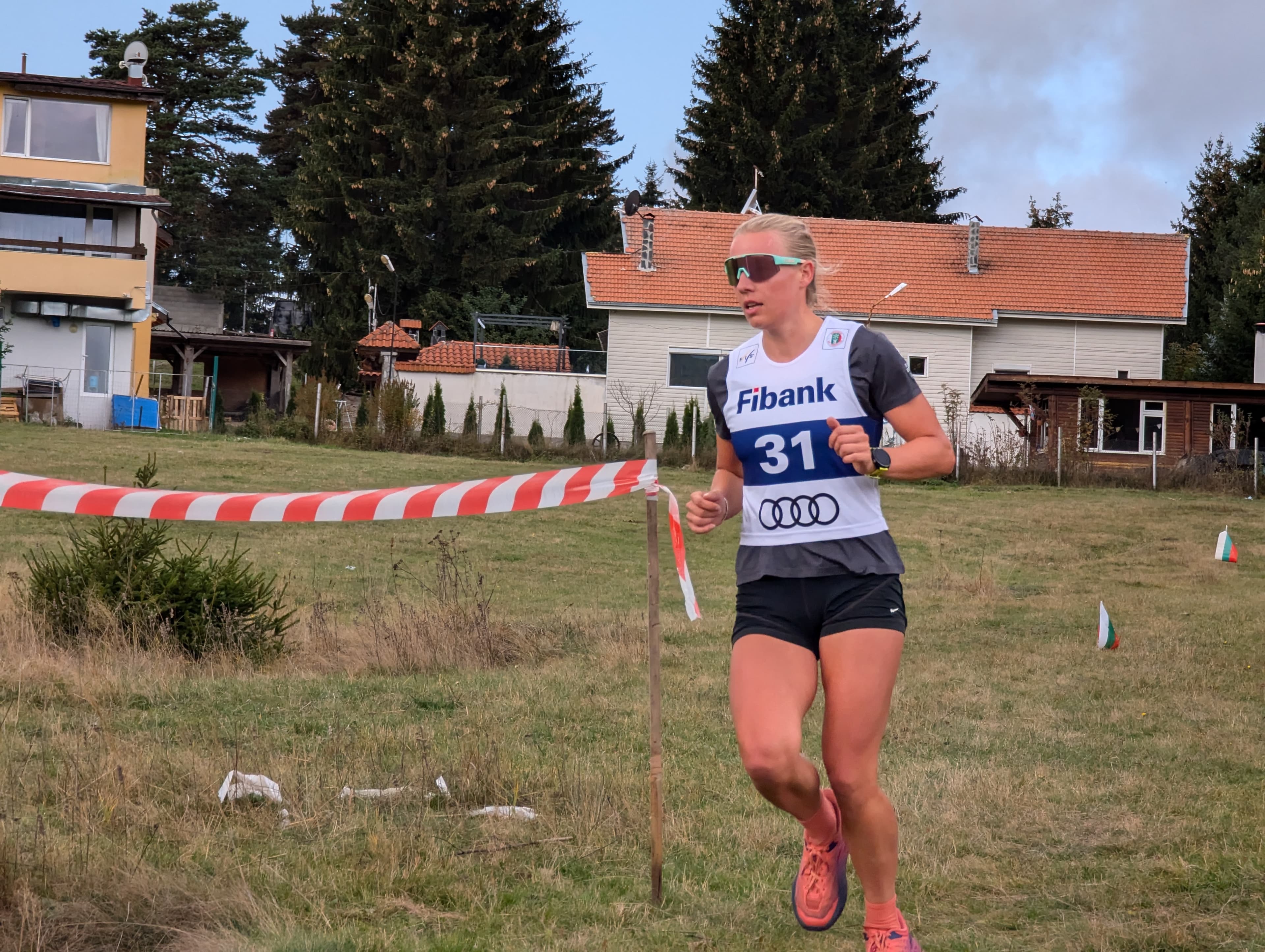A blonde athlete wearing bib 31 runs a cross-country race through a grassy field.