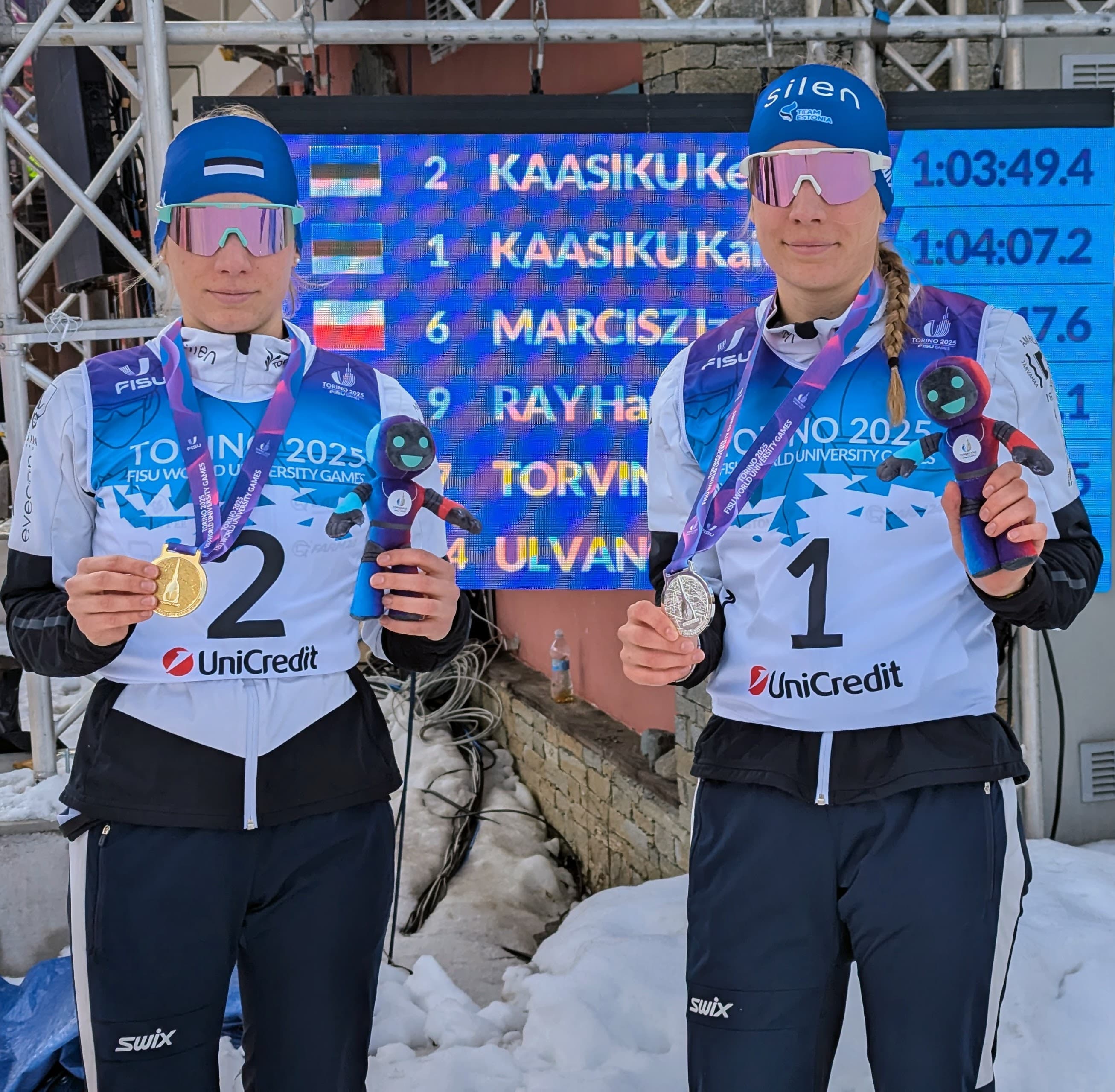 Two Estonian cross-country skiers pose with gold and silver medals and mascots at a scoreboard.