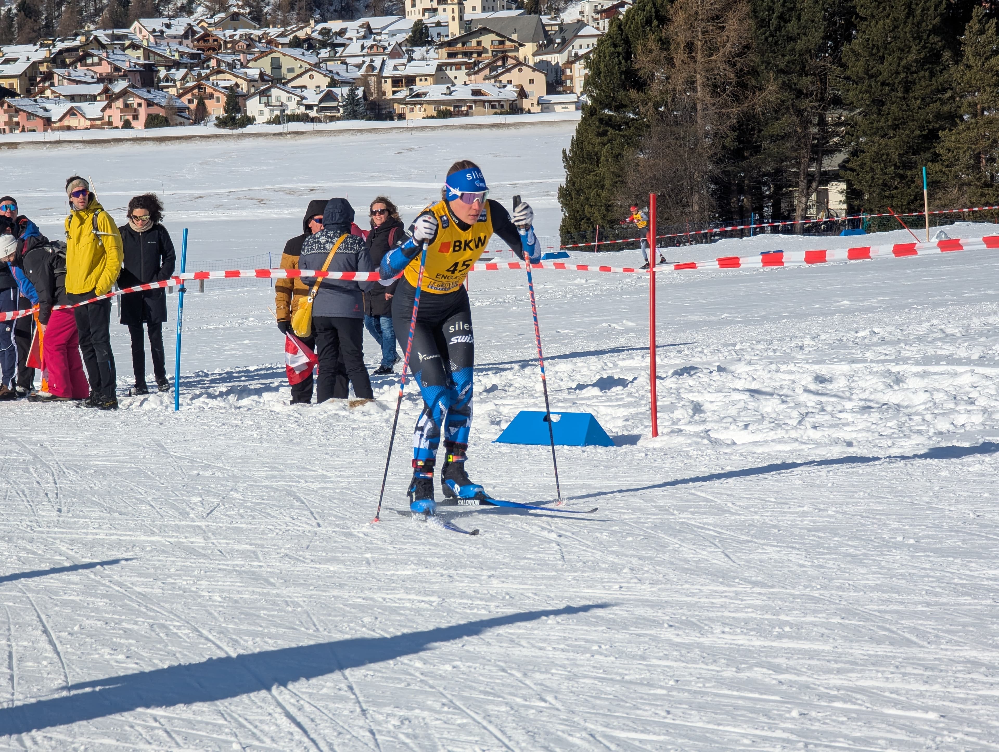 A cross-country skier glides across a snowy track past spectators in a mountain village.