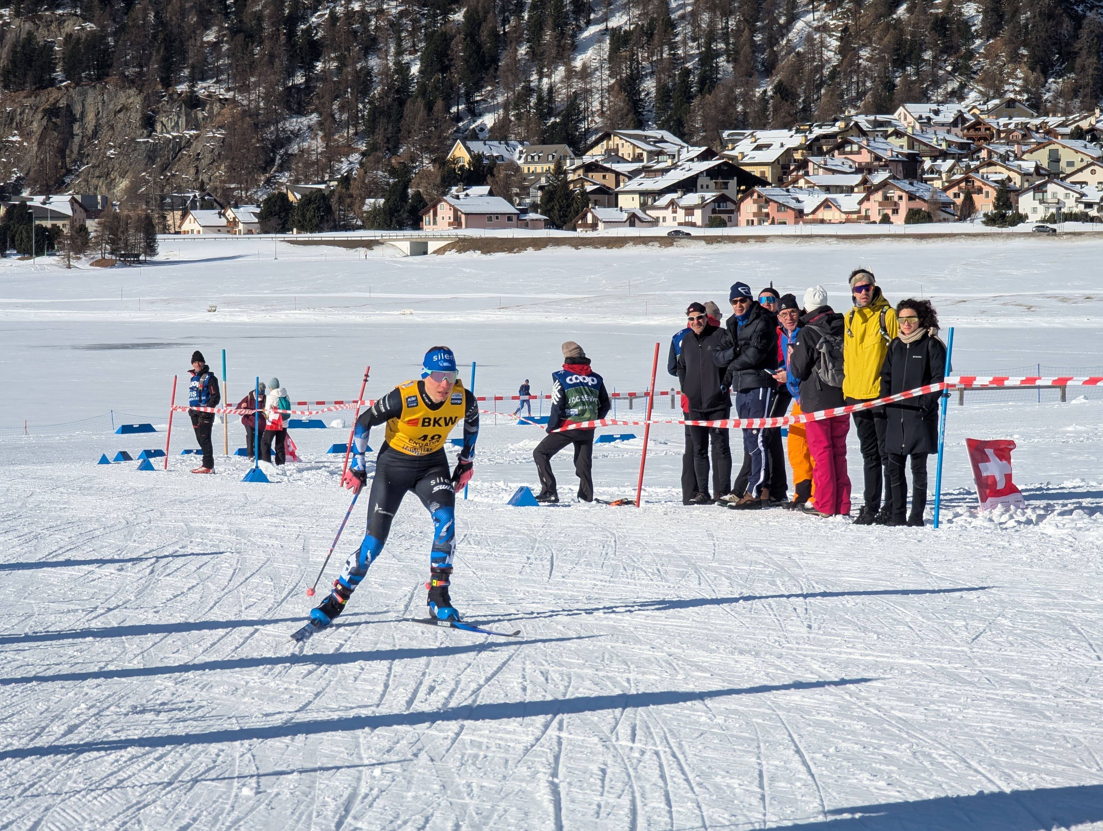 A cross-country skier races across a snowy landscape past spectators and a mountain village.