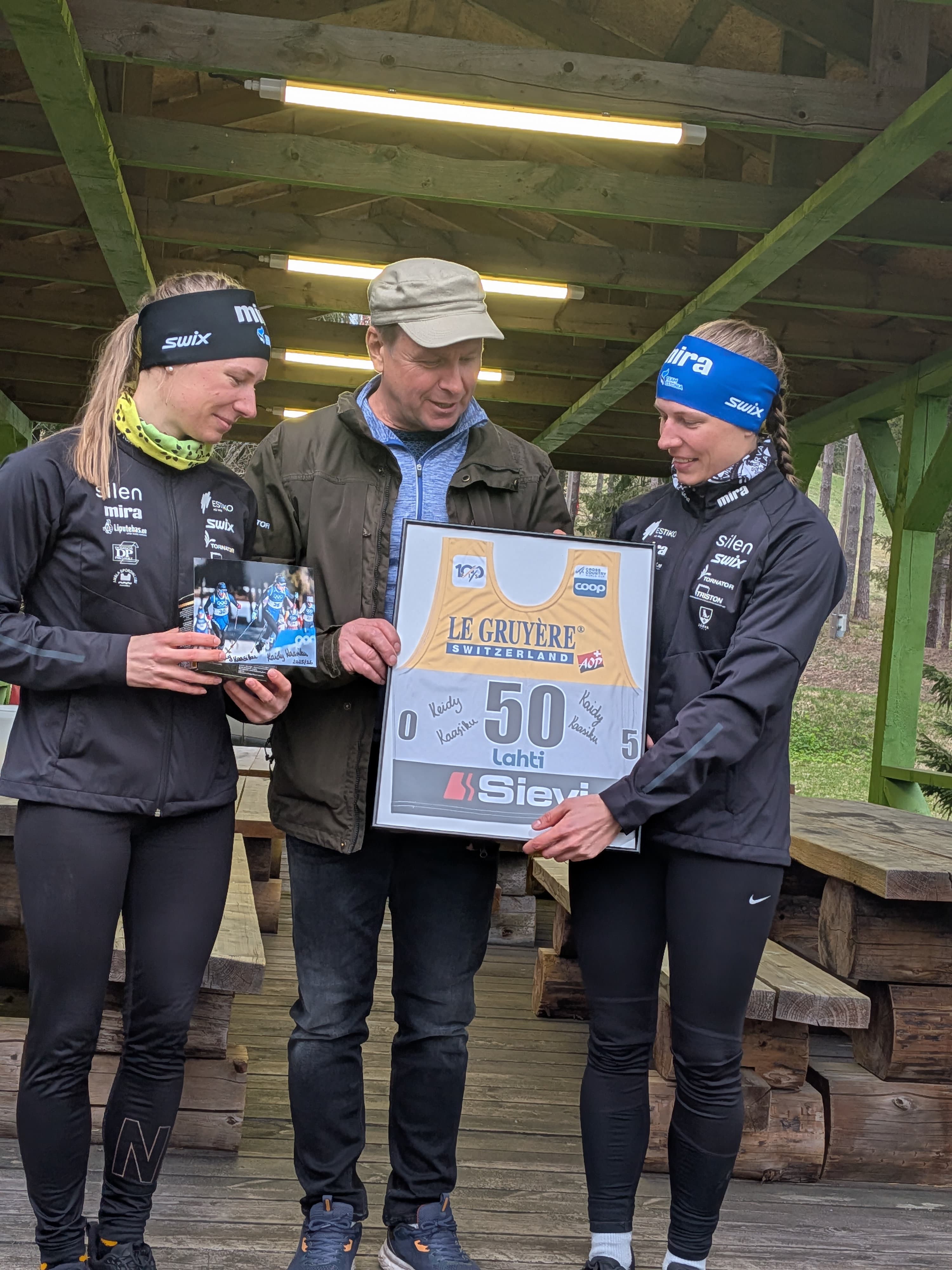 Two athletes and a man holding a framed racing bib and photo under a shelter.
