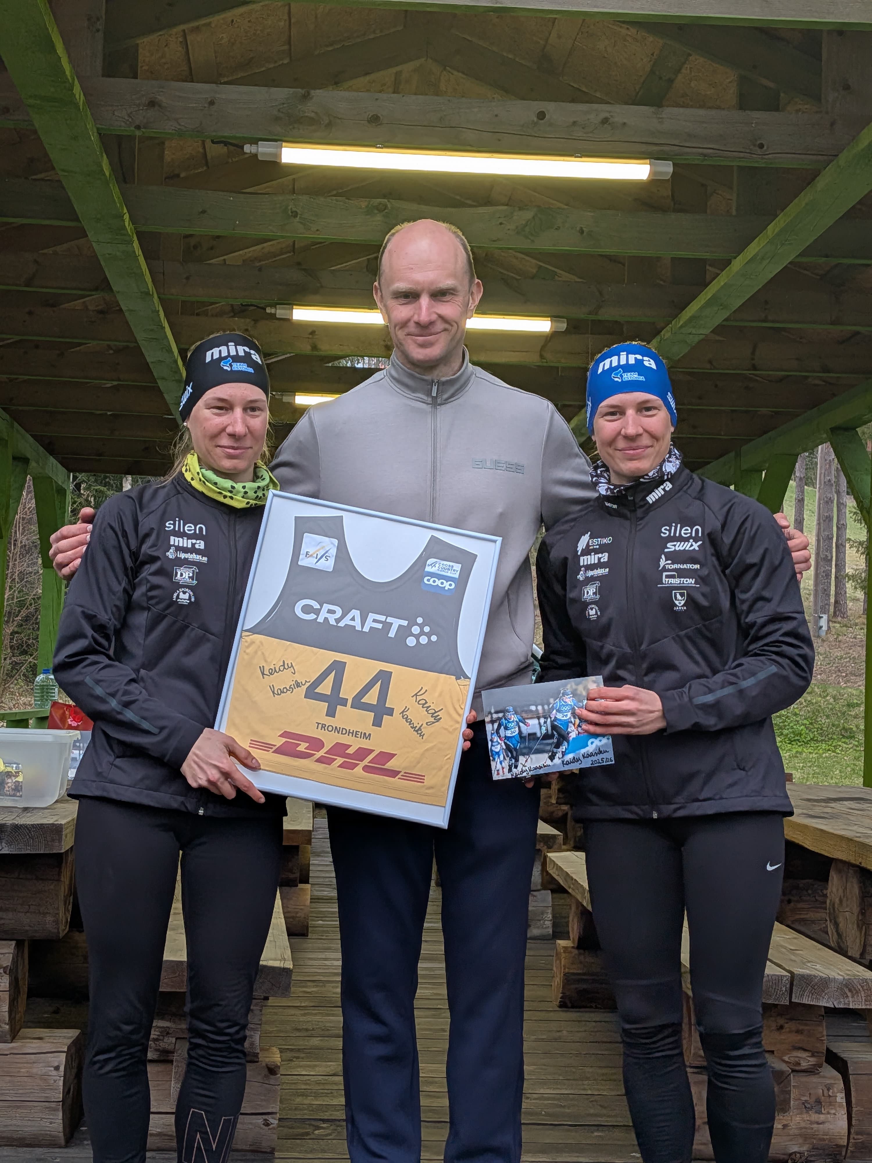 Two female athletes and a man hold a framed racing bib under a wooden shelter.