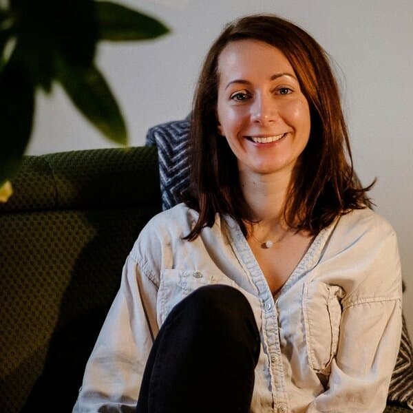 Smiling woman with brown hair wearing a light shirt, sitting on a green couch indoors.