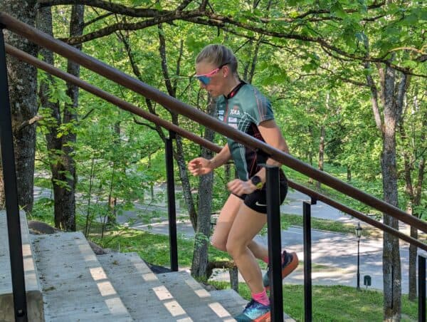 Athletic woman in pink sunglasses runs up concrete stairs through a lush green forest.