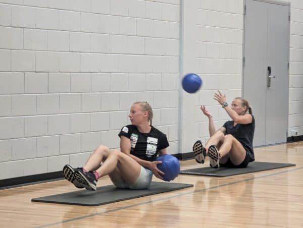 Two women perform seated core exercises on mats while tossing blue medicine balls indoors.