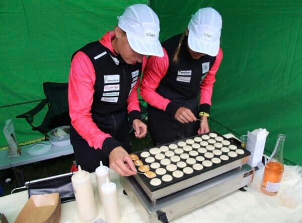 Two women in athletic gear cook small round pancakes on a large griddle outdoors.