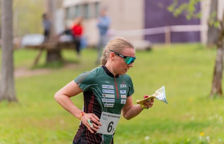 Female orienteer in a green jersey runs through a field while checking a handheld map.