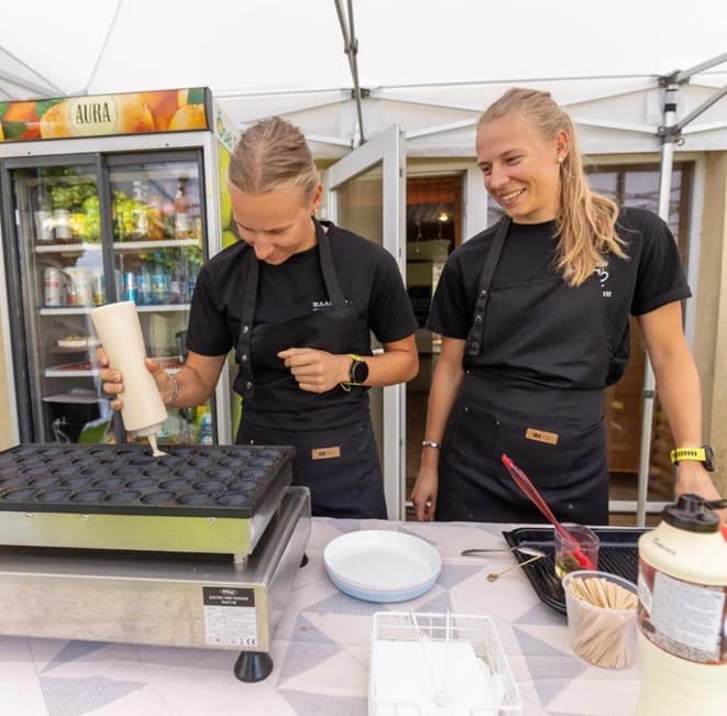 Two women in black aprons prepare mini pancakes at an outdoor food stall.