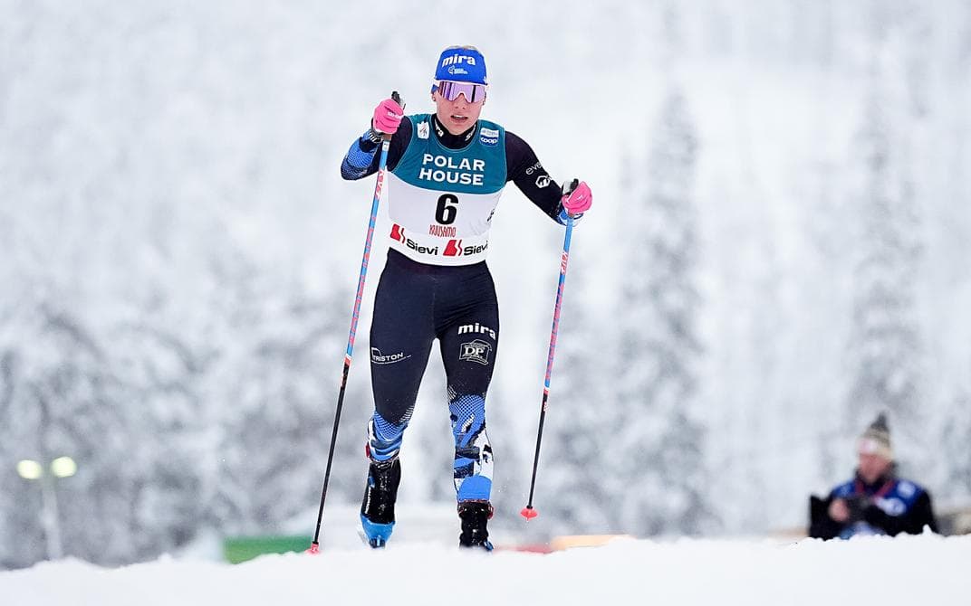 A female cross-country skier wearing bib six glides across a snowy track with poles.